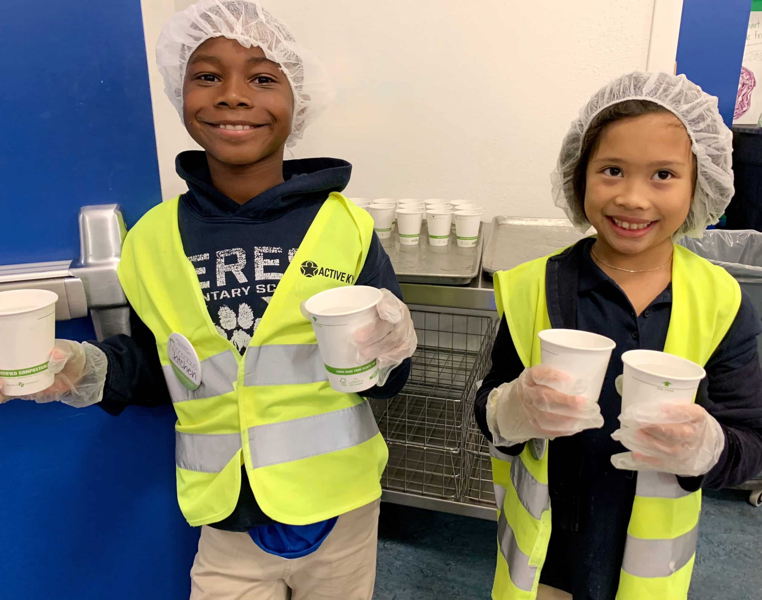 Two students in yellow safety vests, hair nets, and gloves are holding cups of organic milk and smiling at the camera.