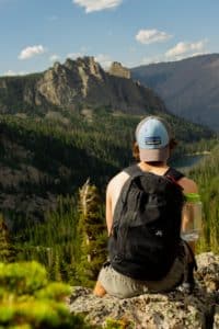 A person in an orange shirt and black backpack, wearing a backwards hat with the Patagonia logo, facing away from the camera and looking out over a scene of mountains during a hike.