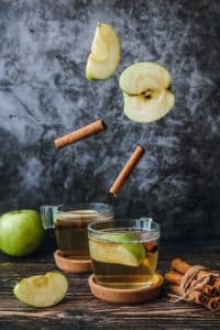A stylized photo of apple cider. There are two short, clear glass mugs on a wooden surface filled with apple cider and a slice of green apple. Above the mugs, as if falling, are cinnamon sticks and more green apple slices. On the surface of the table are more apples and a bunch of cinnamon wrapped with twine.