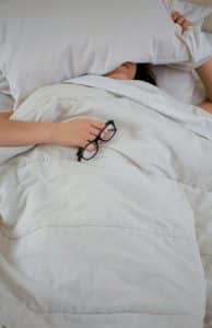 A photo of a woman sleeping, mostly covered by a white comforter and pillow. In her right hand on top of the covers she is holding a pair of black thick-rimmed glasses.