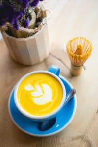 A top down photo of a turmeric latte in a blue coffee cup on a blue saucer. The latte has milk art in the shape of a leaf on top, and the latte itself is a goldenrod color. In the background there is a whisk and a potted flower.
