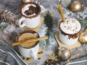 Three mugs of hot chocolate. At top left the cocoa is topped with whipped cream and chocolate sauce. At bottom left the mug is garnished with a cinnamon stick laying across the top of the mug. To the right, the cocoa is topped with whipped cream and shaved chocolate, with a cinnamon stick in the cocoa. The table is dressed with small gold ornaments, pine sprigs, a pinecone, and fake snow.