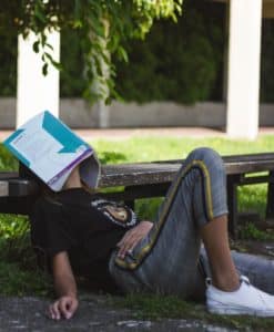 A student rests on the ground, laying against a bench, with a textbook opened over their face.